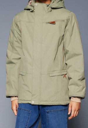 Personne portant une veste à capuche vert clair avec des poches zippées sur le devant et la poitrine, fermeture par boutons et jean en denim bleu.