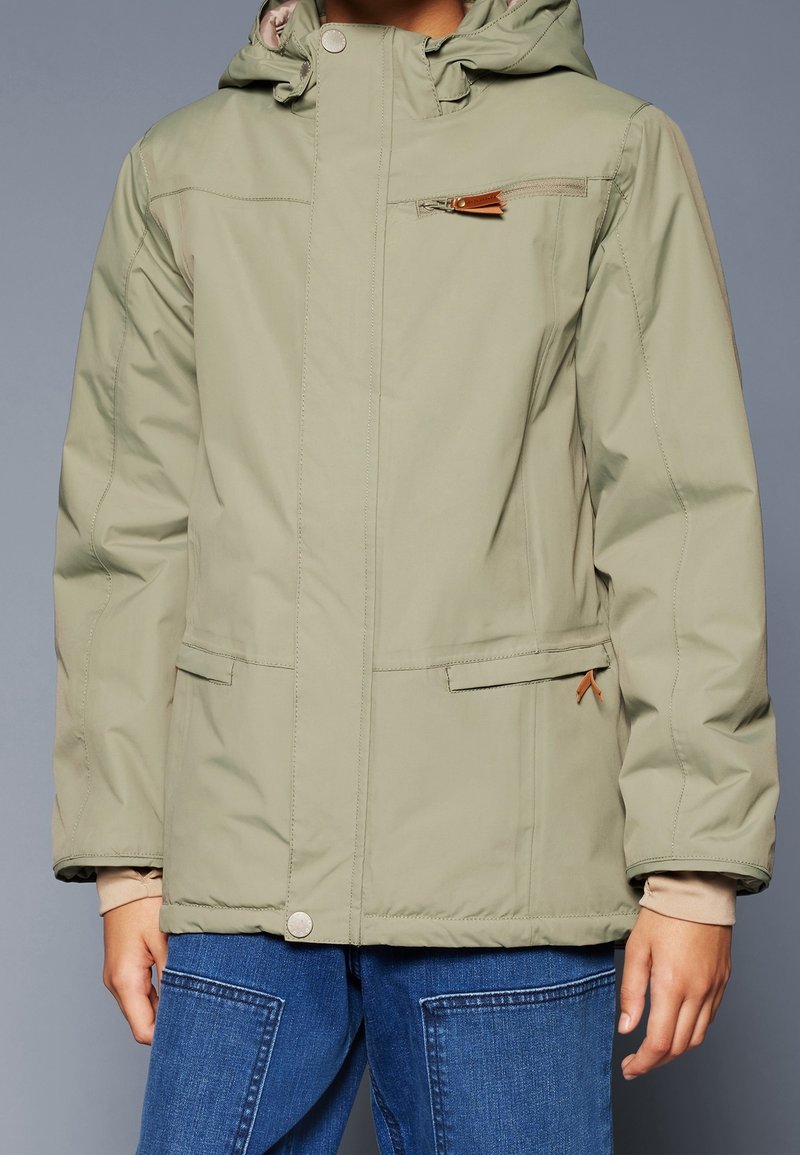 Personne portant une veste à capuche vert clair avec des poches zippées sur le devant et la poitrine, fermeture par boutons et jean en denim bleu.