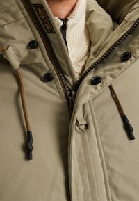 Olivgrön jacka med dragkedja, hög krage, stora knappar och bruna dragsnören. Har ett mjukt, tjockt stickat innerlager.