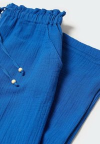 Pantalons texturés bleu avec taille élastique et cordon de serrage, présentant deux perles en bois à chaque extrémité, posés à plat sur une surface blanche.