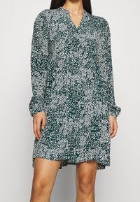 Abito verde con motivo floreale bianco, maniche lunghe, scollo a V e una vestibilità morbida che arriva sopra il ginocchio. Tessuto con texture liscia.