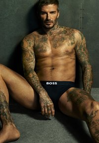 Boxer-briefs noirs avec un logo "BOSS" blanc, mettant en valeur un torse tatoué, une silhouette musclée et des cheveux courts et en pointe sur un fond sombre.