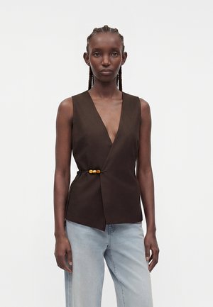 Femme aux cheveux tressés portant un haut cache-cœur sans manches marron foncé avec un fermoir en perles de bois, associé à un jean bleu clair, debout sur fond blanc.