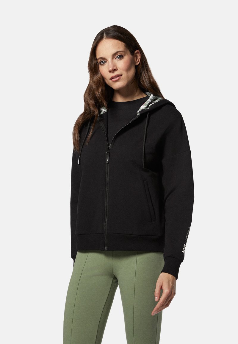 Sudadera negra con cremallera y capucha interior con estampado, presenta un corte holgado, bolsillos y puños acanalados. Combinada con leggings verdes.