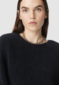 Vrouw met een lichte huid en lang donker haar, gekleed in een donkere fuzzy trui en een chunky zilveren ketting tegen een effen achtergrond.