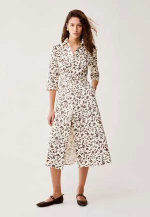 Donna in piedi con una mano in tasca che indossa un vestito camicia midi color crema con stampa floreale marrone e ballerine marroni su sfondo neutro.