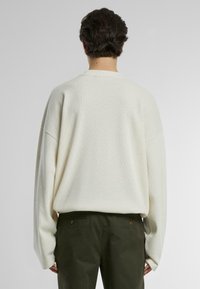 Weißer Strickpullover mit strukturiertem Muster, lockerer Passform und überschnittenen Schultern. Kombiniert mit dunkelgrünen Hosen mit hinteren Taschen.
