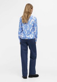 Blå langærmet bluse med hvid abstrakt leopardprint, der har en tekstureret overflade. Sammen med mørkeblå løstsiddende jeans og sorte sko.