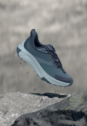 Modrá a sivá atletická topánka vo vzduchu nad skalnatým povrchom s malými úlomkami vznášajúcimi sa okolo, čo naznačuje pohyb alebo skok.