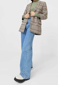 Blazer a doppio petto a tartan beige con accenti rossi e blu, abbinato a jeans a palazzo azzurri chiari e stivali piatti bianchi.
