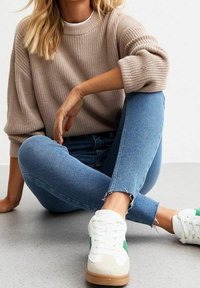 Persona que lleva un suéter de punto beige y jeans skinny azules, sentada con las piernas cruzadas en el suelo, vistiendo zapatillas blancas con una franja verde.