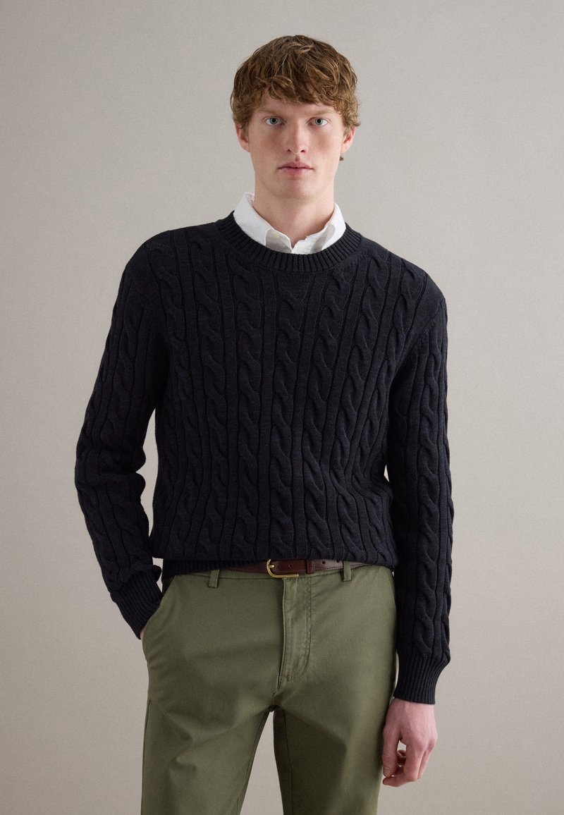 Junger Mann, der einen schwarzen Pullover mit Zopfmuster über einem weißen Hemd und olivgrüne Hose mit braunem Gürtel trägt, vor einem einfarbigen Hintergrund steht.
