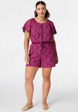 Vrouw met krullend haar, gekleed in een paarse korte mouwen bloemenprint romper en beige gevlochten platte schoenen, staat zelfverzekerd tegen een effen achtergrond.