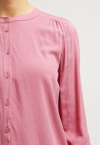 Chemise rose à boutons avec encolure ronde, manches longues et légère fronce à l'épaule ; tissu lisse et patte de boutonnage à six boutons.