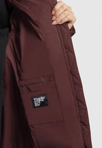 Quiltad maroon jacka med svart etikett på insidan; har en sidficka och en slät, blank tygtextur.