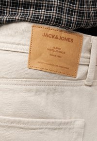 Κοντινή λήψη πίσω τσέπης μπεζ τζιν με καφέ δερμάτινη ετικέτα Jack & Jones και πουκάμισο με ασπρόμαυρο καρό πάνω από αυτή.
