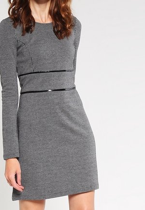 Femme portant une robe gris et noir à manches longues et motif pied-de-poule ajustée, avec une fine ceinture noire à la taille, debout devant un fond neutre.