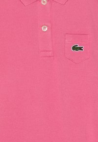 Polo din bumbac roz, cu o țesătură texturată, guler cu trei butoane și un mic logo verde de crocodil pe buzunarul de la piept.
