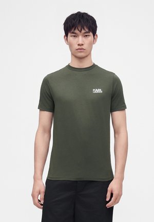 Ung mand med kort sort hår iført en olivengrøn Karl Lagerfeld t-shirt og sorte bukser, stående mod en ensfarvet lys baggrund.