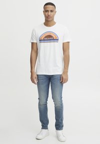 Balta medvilninė marškinėliai su daugiaspalviu grafiniu užrašu "BLEND DENIM ORIGINAL BUILT IN BLUE", derinami su šviesiai mėlynomis džinsomis ir baltais sportbačiais.