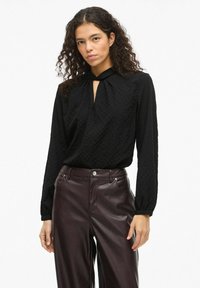 Svart teksturert bluse med nøkkelhullutringning og lange ermer, kombinert med høytlivs burgunderfargede skinnbukser.