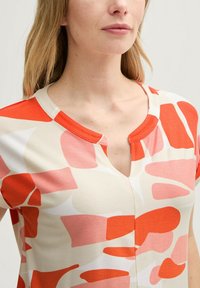 T-shirt à manches courtes avec un col en V, présentant un fond crème et des formes abstraites en corail, rose et blanc. Tissu lisse avec un léger éclat.