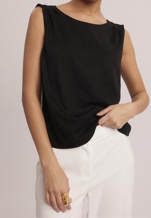 Kobieta w czarnej bluzce bez rękawów i białych spodniach, trzymająca spod bluzki jedną ręką, nosząca dwa masywne złote pierścionki.