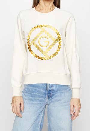 Sudadera blanca con diseño de cuerda dorada y logo; cuenta con mangas raglán y un dobladillo acanalado, combinada con jeans de mezclilla azul claro.