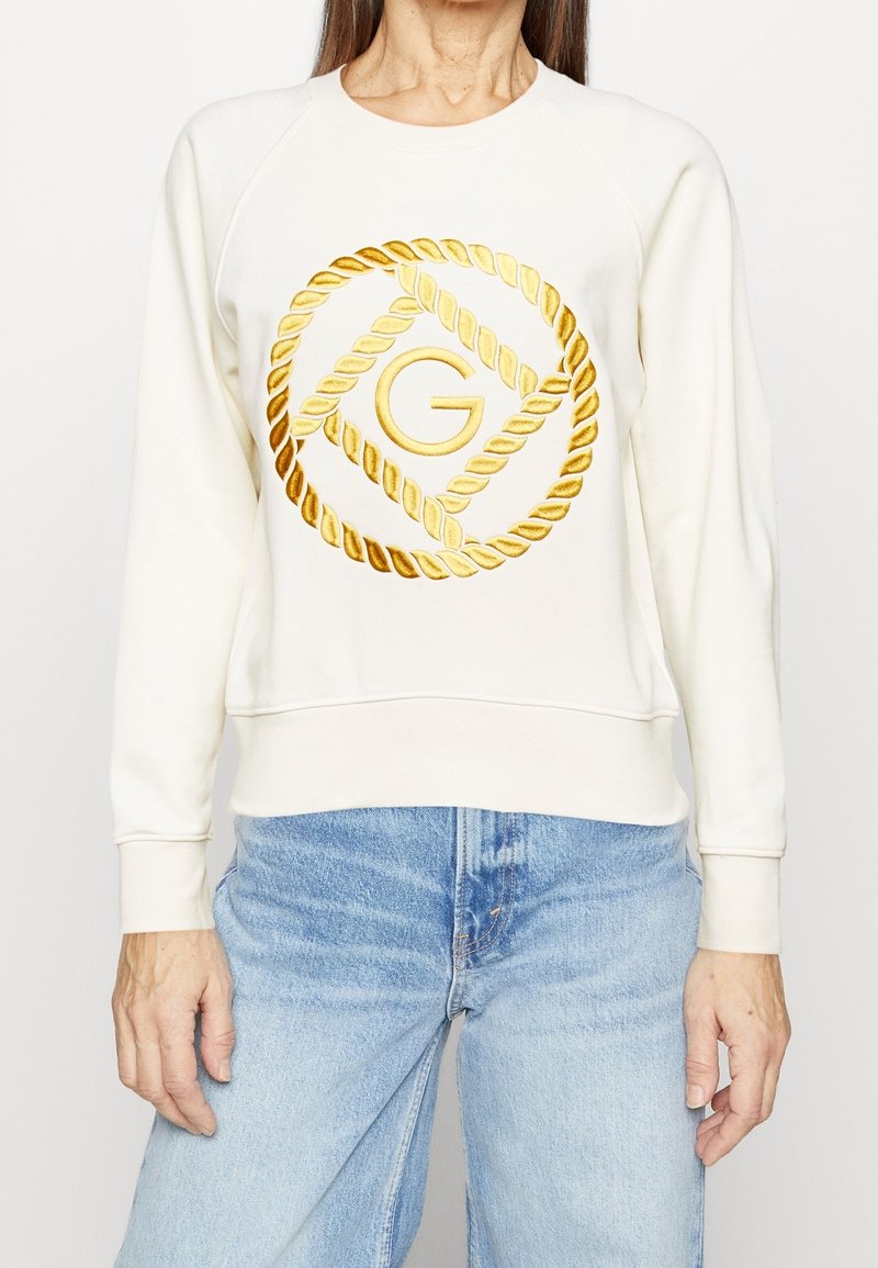 Vit sweatshirt med guldrep och logodesign; har raglanärmar och ribbat avslut, tillsammans med ljusblå denimjeans.