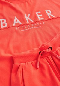 Coral-värinen vapaa-ajan asukokonaisuus, joka sisältää T-paidan, jossa on rohkeaa "BAKER"-tekstiä, sekä yhdistelevät collegehousut, joissa on ribbausvyötärö ja nauhakuji.