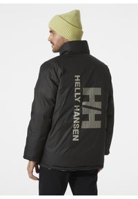 Chaqueta negra acolchada con el logo de Helly Hansen en la parte posterior en gris claro. Complementada con un gorro de punto acanalado en verde claro.
