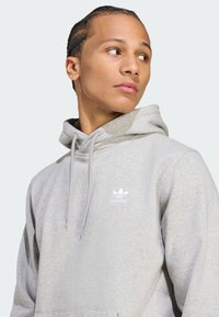 Világosszürke kapucnis pulóver, puha anyagból készült, kenguruzsebbel, húzózsinóros kapucnival és fehér Adidas logóval a mellkason.