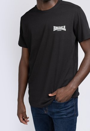 Mann trägt ein schwarzes Lonsdale London T-Shirt mit kurzen Ärmeln und blauen Jeans, Hand in der Tasche vor einfachem weißem Hintergrund.