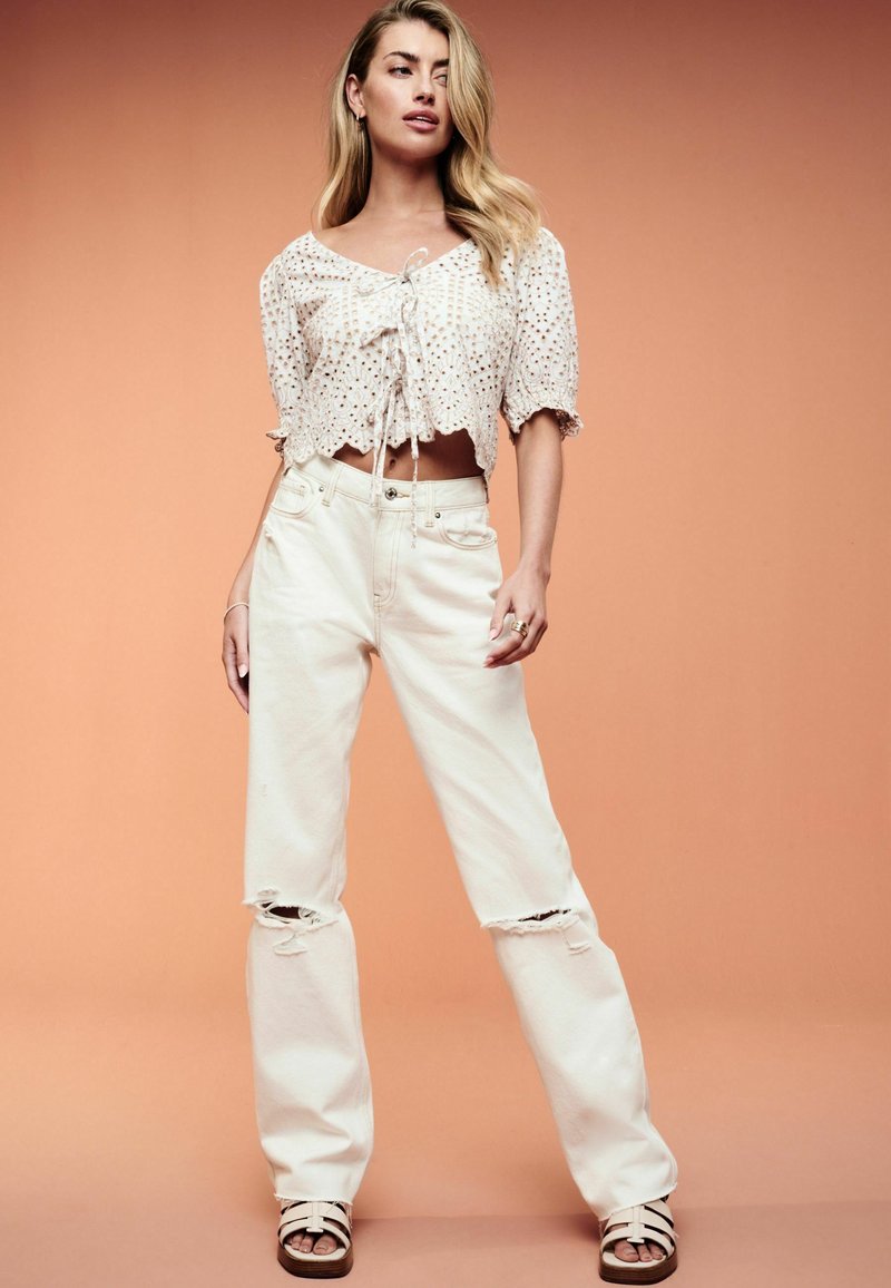 Blusa crema leggermente testurizzata con maniche a palloncino corte e dettaglio con fiocco, abbinata a pantaloni in denim bianco larghi e strappati e sandali.
