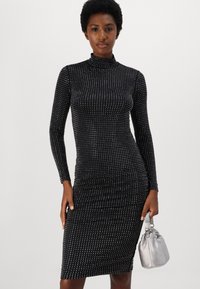 Musta pitkähihaine mekko, jossa on säädettävä siluetti, turtleneck-kaulus ja hopeapistekuvio. Yhdistettynä pieneen hopeiseen käsilaukkuun.