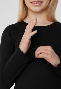 Czarna, prążkowana bluzka z długim rękawem i falbaniastymi mankietami, ręce w pozycji do poprawiania odzieży, uwaga skoncentrowana na detalach rękawów.