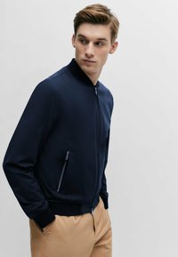 Navy blue bomber jacket with a zip front, ribbed cuffs, and two side zip pockets. Smooth texture with a fitted design and minimal accents.