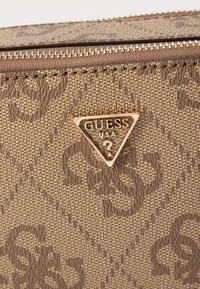 Brązowy teksturowany materiał z wytłoczonym wzorem; posiada złotą trójkątną plakietkę z logo. Zamknięcie na zamek błyskawiczny u góry.