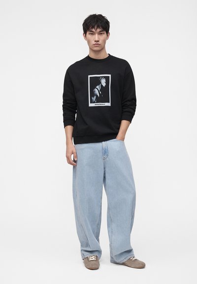 Jeune homme portant un sweat-shirt noir avec un imprimé graphique, un jean large bleu clair et des baskets beiges, debout devant un fond blanc uni.