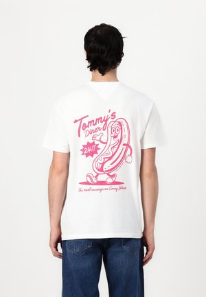 Personne portant un t-shirt blanc avec un hot-dog rose cartoon et le texte « Tommy's Diner 24/7, The best sausage on Coney Island » dans le dos.