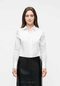 Γυναίκα με ίσια καστανά μαλλιά, ντυμένη με λευκό πουκάμισο με κουμπιά και μαύρη δερμάτινη φούστα, στέκεται μπροστά σε απλό ανοιχτόχρωμο φόντο.