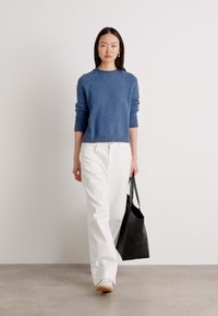 Blauwe gebreide trui met geribde halslijn, gedragen met witte wijde broek. Model houdt een zwarte tote bag vast. Eenvoudig, strak ontwerp.