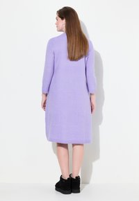 Lila, langärmliges Strickkleid aus weichem Stoff, mit gerader Silhouette und einem Saum, der unter dem Knie endet. Getragen mit schwarzen Stiefeln bis zum Knöchel.