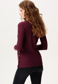 Bordeaux ribbel lange mouwen top met knoopaccenten aan de linkerkant, gecombineerd met zwarte strakke jeans. Glad textuur, casual design.