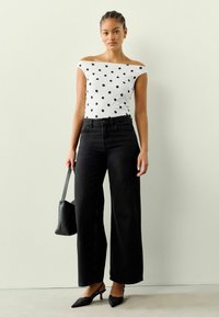 Biała bluzka odkrywająca ramiona w czarne kropki, czarne spodnie z szerokimi nogawkami i czarne buty na obcasie z odkrytymi piętami. Trzyma małą czarną torebkę.