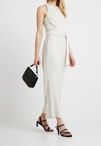 Krämfärgad, ärmlös jumpsuit med vertikala ränder, insvängd midja och vida ben, matchad med svarta remsandaler och en texturerad handväska.