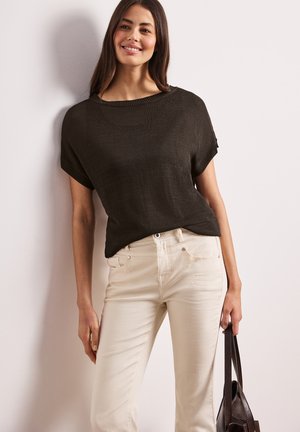 Frau lächelt, trägt ein dunkles kurzärmeliges Strickoberteil und cremefarbene Hosen, hält eine dunkelbraune Handtasche vor einem schlichten hellen Wandhintergrund.