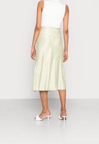 Žena oblečená v světle zelené midi sukni s jemným kostkovaným vzorem, bezrukávovém bílém topu a bílých klínových sandálech z boku.