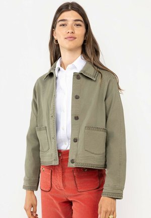 Joven mujer que lleva una chaqueta verde oliva abotonada sobre una camisa blanca y pantalones de tiro alto rojos con bolsillos delanteros, posando contra un fondo blanco.