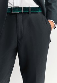 Pantaloni neri su misura con una texture liscia, caratterizzati da una cintura in velluto verde scuro e una fibbia argentata, posizionati in vita con le mani nelle tasche.